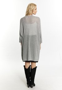 Cardigan in maglia argento con vestibilità ampia, maniche tre quarti e orlo a costine. Indossato sopra un vestito nero e abbinato a stivali neri fino al ginocchio.