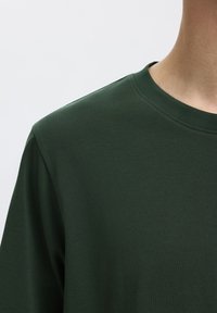 Close-up af en person iført en mørkegrøn t-shirt med rund hals og korte ærmer mod en ensfarvet lys baggrund.