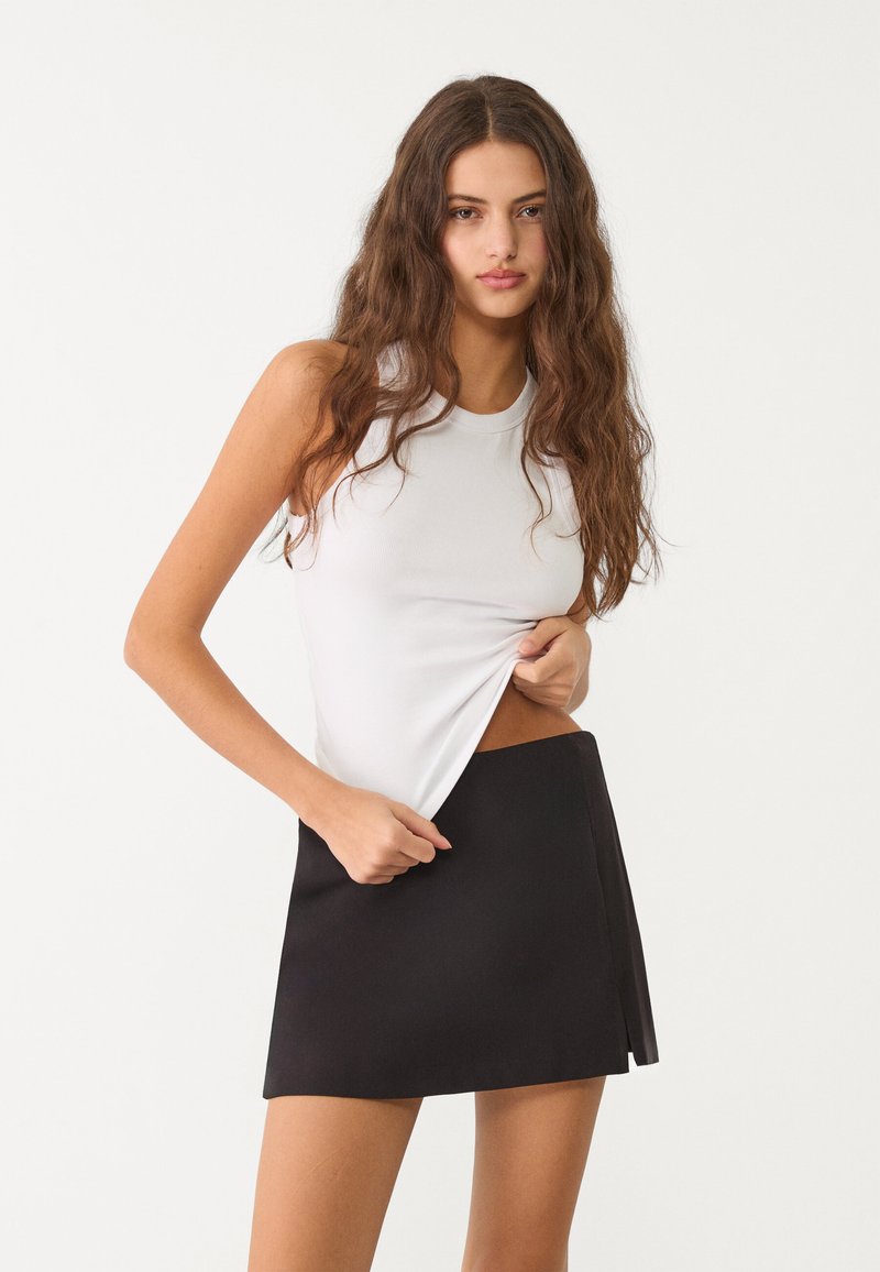 Weißes ärmelloses Top mit gerippter Textur, kombiniert mit einem schwarzen Minirock mit seitlichem Schlitz. Das Outfit hat eine figurbetonte Silhouette.