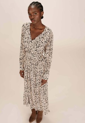 Femme avec des cheveux tressés portant une robe beige à manches longues et col en V avec un imprimé abstrait noir et des bottes brunes à la cheville, se tenant devant un fond clair.