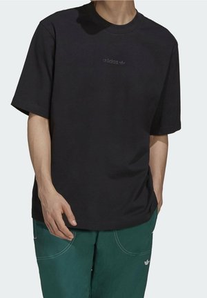 Person iført sort Adidas T-shirt med lille logo på brystet og grønne bukser med hvide syninger og Adidas-logo på lommen.