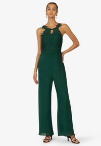 Groene jumpsuit met een bloemenkant lijfje, keyhole halslijn en wijde benen. Strappy zwarte hakken complementeren de outfit.