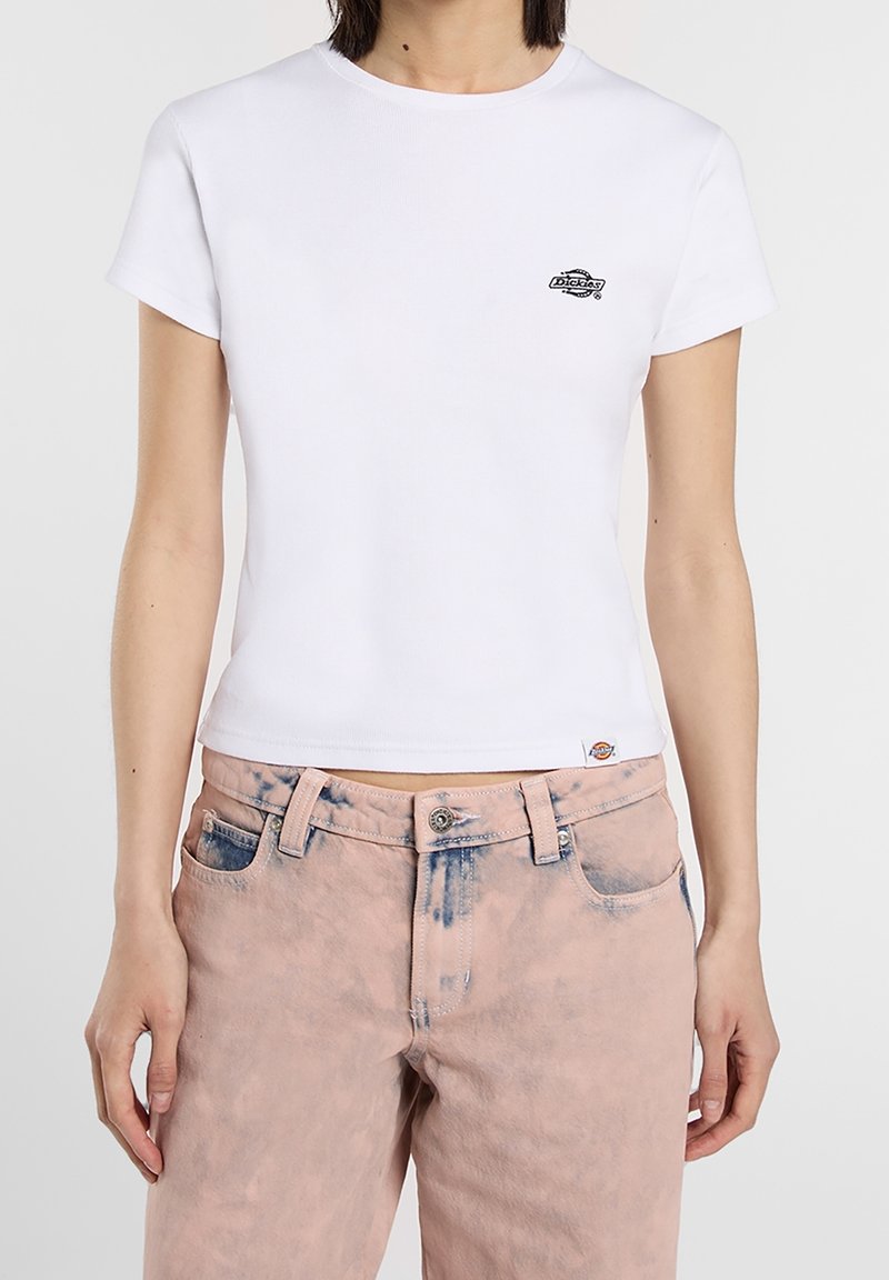 Femme portant un T-shirt blanc ajusté à manches courtes avec un petit logo et un jean rose délavé avec un bouton et des passants de ceinture.