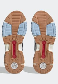 Sulor av sportskor med bruna, ljusblå och vita mönster samt röda torsionsstänger centrerade på varje sulan.