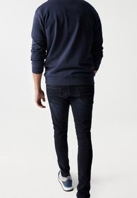 Uomo che se ne va indossando una felpa blu navy, jeans neri e sneakers blu e bianche su uno sfondo bianco.