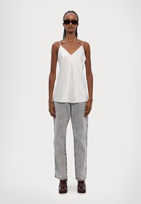 Top blanco sin mangas con escote en V y detalles plisados, combinado con vaqueros rectos gris claro y botas de tobillo marrón oscuro.
