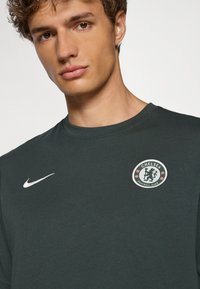 Donkergroene T-shirt met een rond logo van Chelsea Football Club op de linkerborst en twee witte Nike swoosh ontwerpen aan beide zijden.