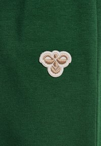 Grön tyg med en strukturerad stickning, med en kräm- och guldbroderad bi-logga på vänster sida. Minimalistisk design, inga ytterligare mönster.