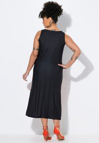 Mustavartaloimainen maksimekko ilman hihoja. Sileä kangas, nilkkapituinen, yhdistetty punaisiin korkokenkiin. Näyttää takaviistosta.