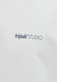 Bílý bavlněný tričko s malým černým logem, které nese nápis "Originals™ STUDIO" v jednoduchém, moderním písmu.