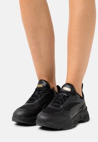 Czarne buty sportowe z teksturowaną cholewką wykonaną z siateczki i skóry, masywną podeszwą oraz złotymi akcentami logo na języku.