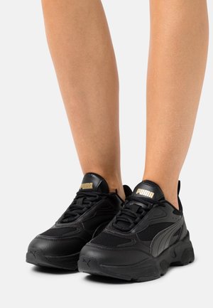 Zapatos deportivos negros con una parte superior texturizada de malla y cuero, suela gruesa y detalles en color dorado del logo en la lengüeta.