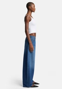Jeans in denim blu a gamba larga con una sfumatura che va dal chiaro allo scuro, caratterizzati da un alto punto vita, tasche posteriori e abbinati a un top corto bianco e scarpe nere.