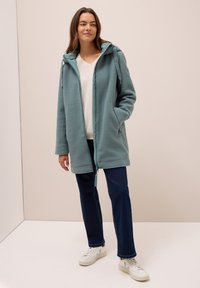 Teal fleece-takki, jossa on vetoketju edessä ja säädettävä huppu. Yhdistetty tummiin farkkuihin ja valkoisiin tennareihin. Pehmeä rakenne ja rento istuvuus.