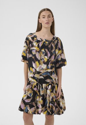 Kobieta ubrana w luźną sukienkę z krótkim rękawem o kolorowym, abstrakcyjnym, kwiatowym wzorze w czarnych, żółtych, różowych, niebieskich i brązowych odcieniach.