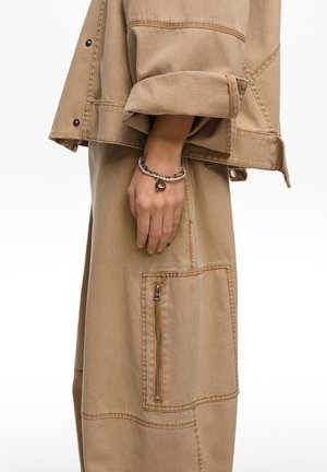 Ręka i przedramię w beżowej kurtce i spódnicy z dużą kieszenią na suwak, ozdobione bransoletką z koralików z serduszkiem jako zawieszką.