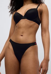 Μαύρο σουτιέν με δαντέλα, με καλυμμένα κύπελλα και ρυθμιζόμενους ιμάντες, συνδυασμένο με μαύρο βαμβακερό στρινγκ που φέρει κόκκινη λεπτομέρεια λογότυπου στη ζώνη.