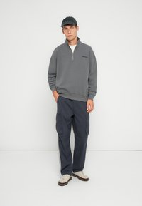 Sudadera de forro polar gris con media cremallera y logo bordado, combinada con pantalones cargo azul oscuro con bolsillos laterales y rayas laterales en contraste, y zapatillas beige.