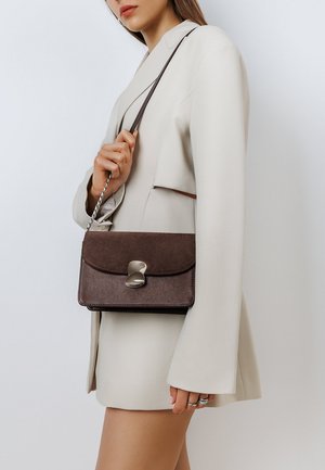 Brown leather handbag with a smooth finish, rectangular shape, flap closure, and metallic hardware. Worn with a light-colored blazer.