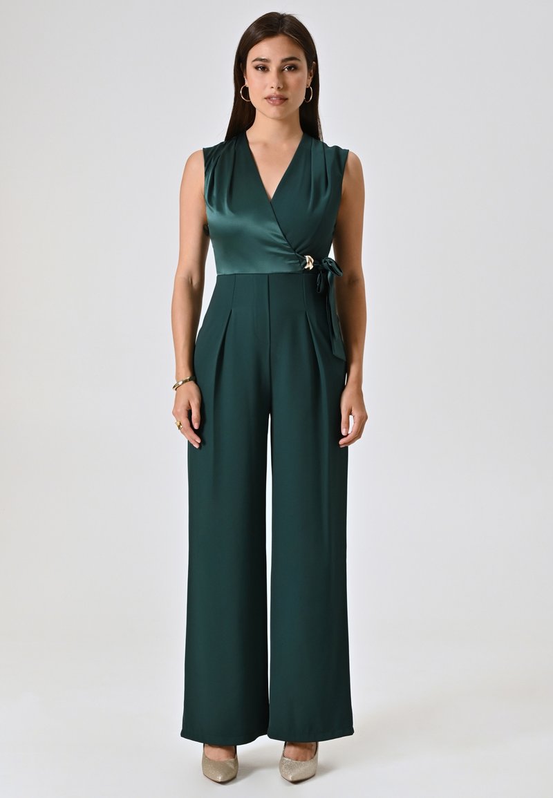Groene satijnen jumpsuit met een wikkelontwerp, mouwloze stijl, geplooide wijde benen en een gestrikt tailleaccent. Beige hoge hakken maken de look compleet.