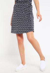 Kvinde iført en sort-hvid mønstret nederdel med et bælte og hvide slip-on platform sko, poserer med det ene ben let hævet.