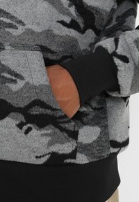 Suéter de camuflaje gris con textura suave, que presenta un bolsillo frontal y puños acanalados negros. El diseño incluye diferentes tonos de gris y negro.