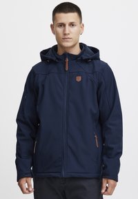 Veste softshell bleu marine avec fermeture éclair à l'avant, capuche ajustable, poches latérales et accents en cuir marron. Textures légère et lisse.