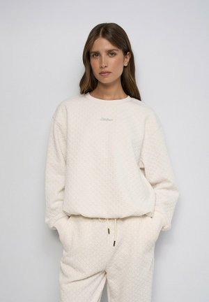 Femme aux cheveux bruns portant un sweat-shirt écru texturé et un pantalon assorti, les mains dans les poches, sur un fond clair uni.
