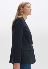 Blazer bleu marine avec une coupe ajustée, fermeture à un bouton, deux poches avant et une texture lisse. Design simple avec des lignes épurées.