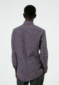 Långärmad skjorta med lila blommönster, knappknäppning vid kragen och rundad nederkant. Tyget verkar lätt och har en något texturerad yta.