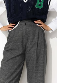 Pantalons à carreaux gris avec taille haute, plis et poches latérales, associés à un gilet en maille bleu marine avec un accent en forme de lettre verte.