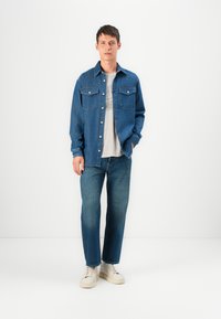 Chaqueta de mezclilla con dos bolsillos en el pecho, cierre de botones y color azul claro, llevada sobre una camiseta gris claro y combinada con jeans azul oscuro.