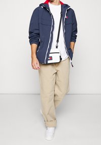 Marinblå vindjacka med röda accenter, vit dragkedja och två framfickor. I kombination med en vit t-shirt, beige byxor och vita sneakers.