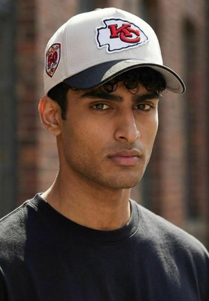 Junger Mann mit lockigem Haar, der eine weiße Kansas City Chiefs Kappe und ein schwarzes Hemd trägt, steht im Freien in der Nähe einer verschwommenen Backsteinwand.