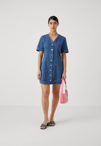Vestido camisola em denim com mangas curtas, abertura frontal de botões e decote em v. Apresenta botões prateados e costura em contraste. Combinado com uma mala de ombro rosa.