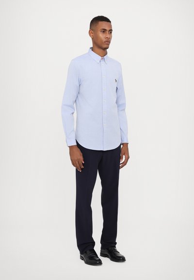 Chemise boutonnière bleu clair avec un col, manches longues et un petit logo sur la poitrine ; associée à un pantalon bleu marine et des chaussures noires.