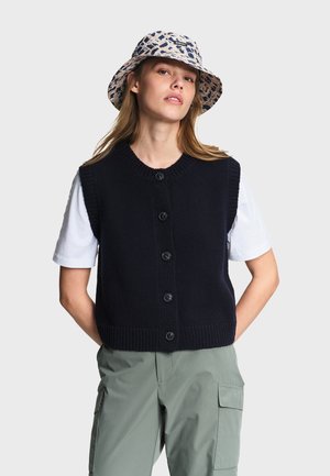 Frau trägt einen gemusterten Anglerhut, eine schwarze, geknöpfte Strickweste über einem weißen Kurzarmshirt und grüne Cargohosen, steht mit den Händen hinter dem Rücken.