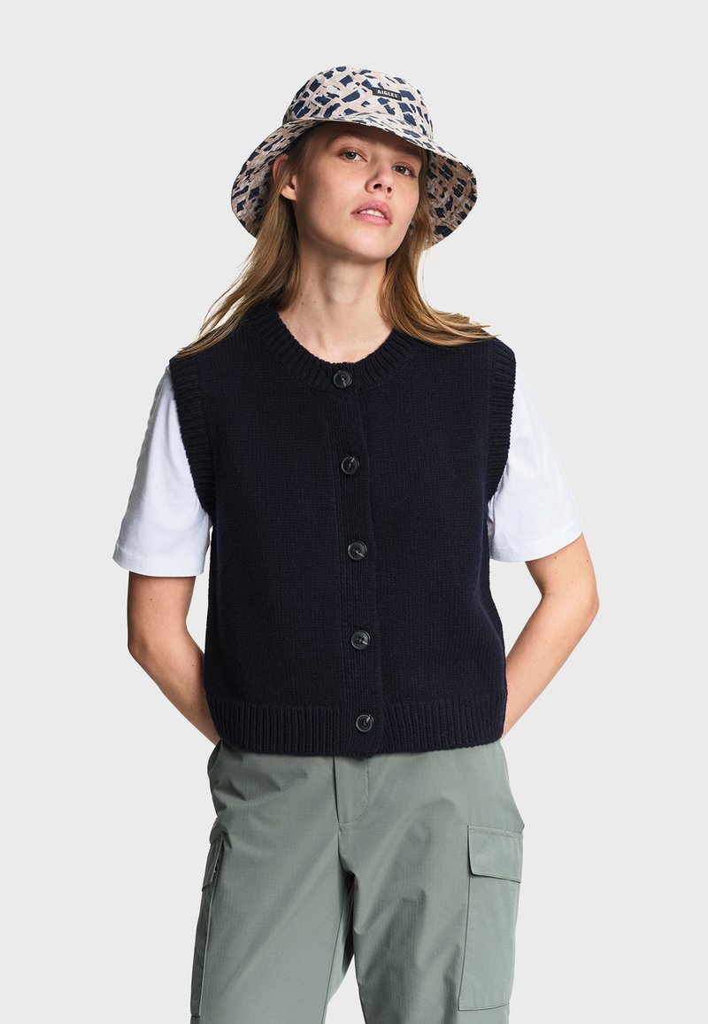 Femme portant un chapeau bob à motifs, un gilet en maille noir boutonné sur une chemise blanche à manches courtes, et un pantalon cargo vert, debout avec les mains derrière le dos.