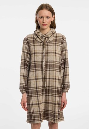 Frau trägt ein beige-braunes kariertes langärmeliges Kleid mit einem gerafften Kragen mit Kordelzug, steht vor einem schlichten weißen Hintergrund.