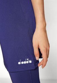 Vijolične atletske kratke hlače iz dihamočega materiala, ki imajo gladko teksturo in bel logo "diadora" na robu.