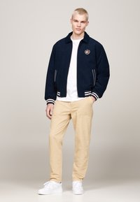 Giubbotto bomber blu con polsini e colletto a righe bianche, decorato con un logo sul petto, abbinato a chinos beige e sneakers bianche.
