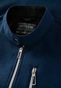 Blå, texturerad jacka med hög krage och vita dragkedjor. Inuti finns märkesetiketten "ombre quintessence". Finns i medelstorlek.