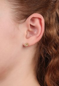 Orecchini a perno in tonalità oro con design circolare, superficie texture e piccoli accenti decorativi, posizionati su un tono di pelle chiara.