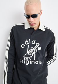 Cămașă neagră cu mâneci lungi, guler alb și trei dungi albe pe mâneci, având un grafic cu un cipit și textul "adidas originals".
