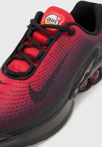 Basket rouge et noire avec une tige en mesh, présentant un dégradé texturé, des lacets et un amorti d'air visible dans la semelle. Marque Air Max visible.