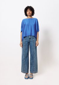 Blause Seidenbluse mit lockerer Passform, kombiniert mit weit geschnittenen, hellblauen Jeans aus Denim mit vorderen Nähten. Blaue Slipper runden das Outfit ab.