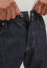 Jeans de mezclilla azul oscuro con cierre de botón y cremalleras, que presentan costuras amarillas y un diseño de cinco bolsillos. Unas manos agarran la cintura.