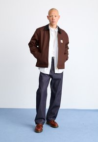 Brown jacket with a black collar, light shirt underneath, dark denim pants, and brown shoes. Simple design with a front zipper and logo patch.