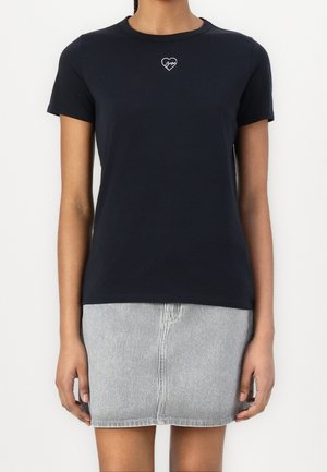 Kvinde iført en tætsiddende sort T-shirt med korte ærmer med et lille hvidt hjerte og signaturlogo på brystet, kombineret med et lysstripet skørt.