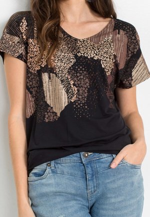 Nainen pukeutuneena lyhythihaiseen, V-kaula-aukkoinen mustaan paitaan, jossa on abstrakteja beigejä ja ruskeita kuvioita, yhdistettynä vaaleansinisiin farkkuihin, käsi taskussa.